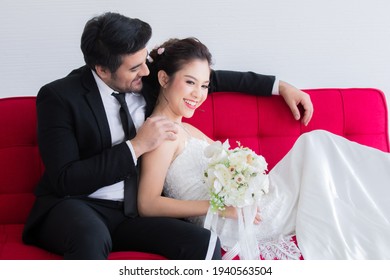 Mix Race Wedding Concept, Beautiful Asian Woman Lie Down On Chest Of Caucasian Handsome Man On Red Sofa Bed, Happy Couple Spent Time Together, Man Taking Care Bride With Love And Tenderness