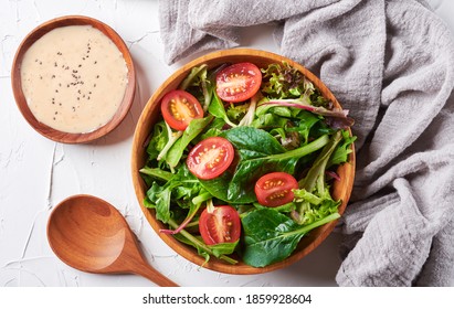 mix salad from green arugula leaves and cherry tomato in wooden bowl and spoon with salad sauce on white stone background. top view - Powered by Shutterstock