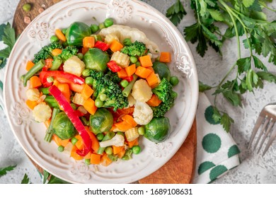 Mix Of Stewed Vegetables In A Ceramic Plate On A Served Table, Top View. Boiled Brussels Sprouts, Carrots, Broccoli, Peas, Peppers And Corn. Cooked Vegan Or Vegetarian Food.