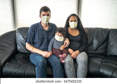 A mixed family sits on a black leather sofa with father and mother holding their infant son all wearing medical face masks in hopes of preventing getting sick from caronavirus or COVID-19.