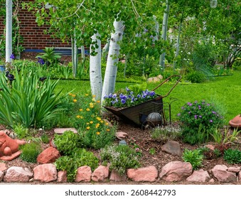 A mixed garden setting bordered by rocks with decorative elements including a wheelbarrow filled with pansies, a variety of flowers herbs and succulents and a Quacking Aspen Tree.