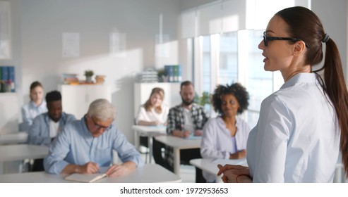 Mixed Group Of Students Listening To Tutor In Class. Young Woman Professor Lecturing In Classroom For Adult People Attending Post Graduate Education Courses In College