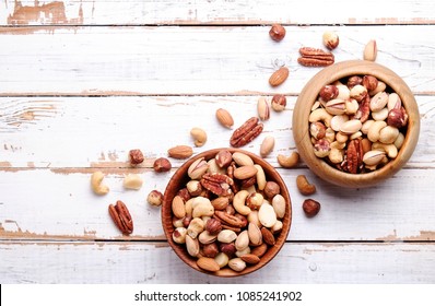 Mixed Nuts In Wooden Bowl And Scattered On Table. Trail Mix Of Pecan, Almond, Macadamia & Brazil Edible Nuts With Walnut Hazelnut On Wood Textured Surface. Background, Copy Space, Top View, Close Up.