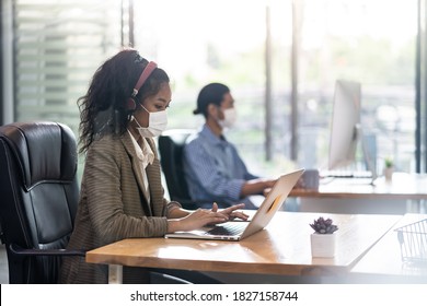 Mixed Race Of African Black And Asian Businesswoman Office Worker Wear Face Mask Working In New Normal Office With Social Distance To Colleague In Background Prevent Coronavirus COVID-19 Spreading.