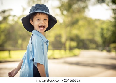 Mixed Race Asian Caucasian Boy Confidently Leaves Home On His First Day Of School. Wearing Uniform And Sun Hat. Walking Down His Suburban Street In The Summer Sun