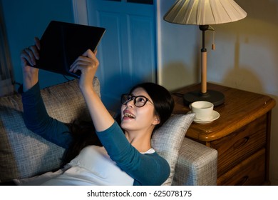 Mixed Race Asian Chinese Student Using Big Digital Tablet Computer Lying Down On The Sofa Facetime With Family At Night When Studying In The Overseas Everyday.