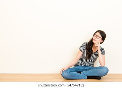 Mixed Race Asian Woman Thinking Question Feel Confusion. Wearing T-shirt Top Sitting On Wooden Floor Over Blank Copy Space White Wall Background.