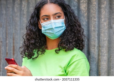 Mixed Race Biracial African American Teenager Teen Girl Young Woman Wearing A Face Mask And Using Mobile Cell Phone For Social Media During The Coronavirus COVID-19 Pandemic