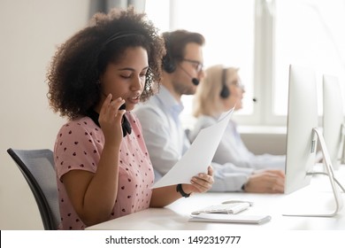 Mixed race call center operator in wireless headset holding, reading paper documents, sitting at workplace in customer support service office, woman in headphones consulting client on phones