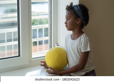 Mixed Race Child With A Soccer Ball In Her Hands Is Sad In The Room. School-age Girl Looks Out The Window Alone. Afro Hair. Concept Of Communication Deficit During A Pandemic, Loneliness In Childhood.