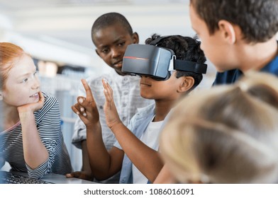 Mixed Race Child With Vr Virtual Reality Goggles In Classroom. Multiethnic Pupil Having Fun With Virtual Reality Headset At Elementary School. Happy Boy Gesturing While Using VR Headset In Classroom.