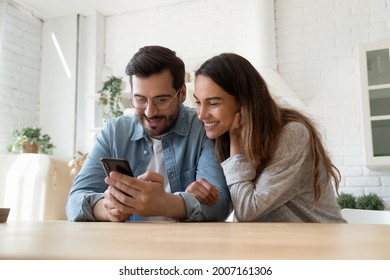 Mixed race couple sit in kitchen at table looking at smartphone screen interested with good e-commerce offer, sale low price and discount in retail services, buying ticket. New mobile app, fun concept