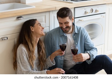 Mixed race couple sit on floor drink red wine relieving fatigue at moving day, celebrate relocation to own house. Romantic dating, kitchen renovation services ad, relations, housewarming event concept