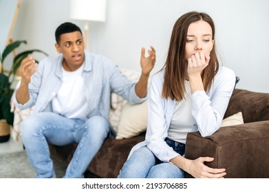 Mixed Race Couple Sitting On A Sofa In The Living Room, An Angry Man Yells At A Woman, Feels Angry, Frustrated Sad Girl Turned Away From Him. Misunderstanding In Relationships, Jealousy, Quarrel