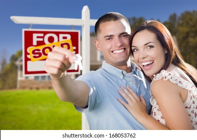 Mixed Race Excited Military Couple In Front Of New Home With New House Keys And Sold Real Estate Sign Outside.