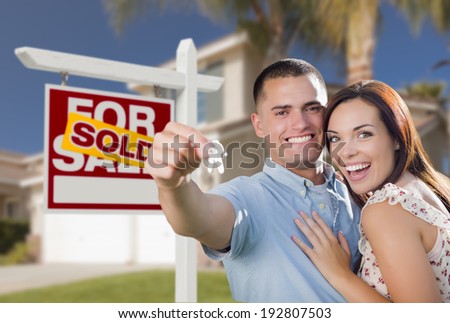 Mixed Race Excited Military Couple In Front of New Home with New House Keys and Sold Real Estate Sign Outside.