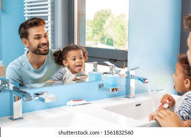 Mixed race family father and little daughter brushing teeth together looking at mirror reflection showing tongue playful smiling cheerful
