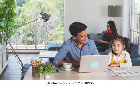 Mixed race family staying together, Caucasian father sitting and working at desk and teaching half-race little cute daughter while Asian mother using laptop computer on sofa. Idea for work at home.
