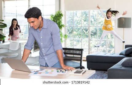 Mixed Race Family Staying Together, Caucasian Dad Working At Home At Desk While Mother Prepare Food And Drink In Kitchen And Little Daughter Play And Jumping On Sofa In Blur, Idea For Warm Family.