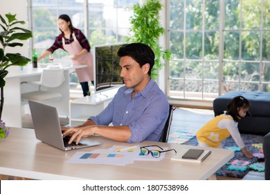 Mixed Race Family Staying Together, Caucasian Dad Working At Home At Desk While Mother Prepare Food And Drink In Kitchen And Little Daughter Play Beside Sofa In Blur Background, Idea For Warm Family.