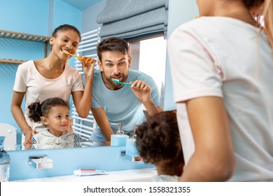 Mixed Race Family Of Three Mother Father And Daughter At Bathroom Brushing Teeth Looking At Mirror Reflection Smiling Playful