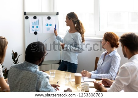 Mixed race female business coach presenting report standing near whiteboard pointing on sales statistic shown on diagram and chart teach diverse company members gathered together in conference room