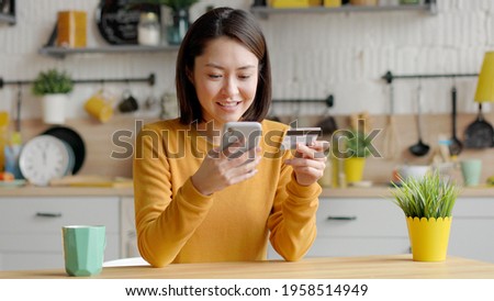 Mixed race female client holding banking credit card and smart phone involved in verification process in mobile application. Asian happy young woman doing payments online or shopping in internet store
