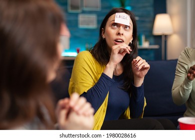 Mixed race friends with sticky notes attached on forehead playing guess who game during home party. Multi-ethnic people having fun, laughing together while sitting on sofa in living room late at night - Powered by Shutterstock