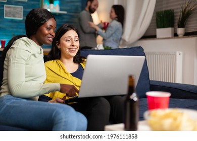 Mixed Race Friends Talking While Looking At Funny Video Series On Laptop Sitting On Couch In Living Room. In Background Woman And Man Drinking Beer Enjoying Time Together During Fun Party.