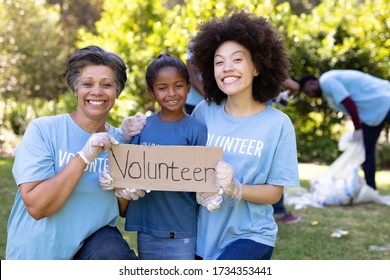 Mixed Race Girl Spending Time Outside With Her Family, Holding A Volunteer Sign With Her Mother And Grandmother, Looking At The Camera And Smiling, All Wearing Blue Volunteer T Shirts, On A Sunny Day