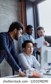 Mixed Race Group Of Colleagues Talking In Friendly Manner Standing Around Asian Man Working On Computer, Business People Enjoy Mutual Communication.