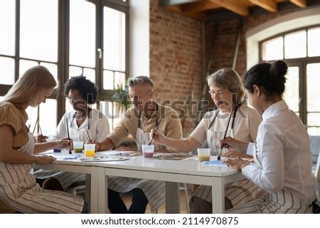 Mixed race group of different aged students drawing on art school class, painting at table, mixing colors, blending paints on palettes, enjoying artistic hobby together, talking, chatting