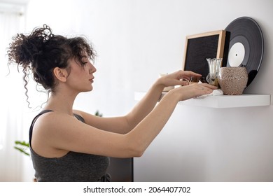 Mixed Race Housewife Cleaning Shelf With Hygge Decor Elements Vinyl Plate Vase Picture Dust Off With Rag. Adorable Curly Female Clean Wiping Wooden Surface Furniture At Home Living Room Apartment