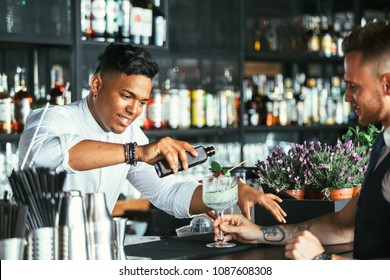 Mixed Race Male Expert Bartender Is Serving Some Tonic For A Cocktail At The Bar Counter While A Smiling Waiter Is Holding The Cocktail Glass