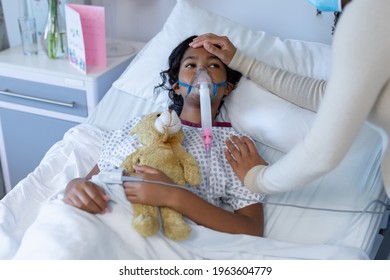 Mixed race mother in face mask touching sick daughter's head on oxygen ventilator in hospital. medicine, health and healthcare services during covid 19 coronavirus pandemic.