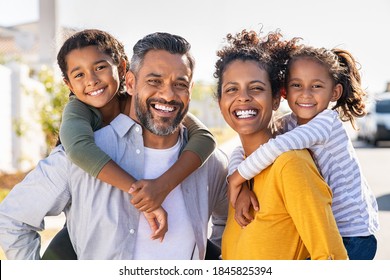 Mixed race parents giving piggyback ride to their children. Portrait of happy african mother and indian father with daughters looking at camera. Smiling family standing with their little girls.