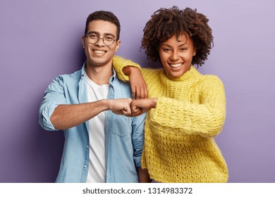 Mixed race partners give fist bump, agree to work together, have happy facial expressions rejoice success and cooperation, have toothy smiles, isolated over purple background. Team work concept