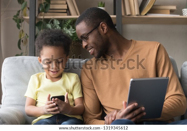 家庭のソファに置かれた道具を使った混血児の息子と黒人の父親 楽しいデジタルタブレット電話を持つかわいい子どもとのアフリカの父親 家族機器の技術コンセプト の写真素材 今すぐ編集