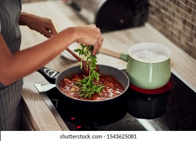 Mixed Race Woman In Apron Standing Next To Stove, Stirring Tomato Sauce And Adding Rocket. On Stove Is Pot With Boiling Pasta.