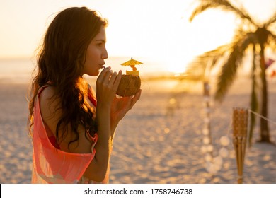 Mixed race woman enjoying her time at a beach with her freinds during sunset, drinking a drink from a coconut, looking away and smiling