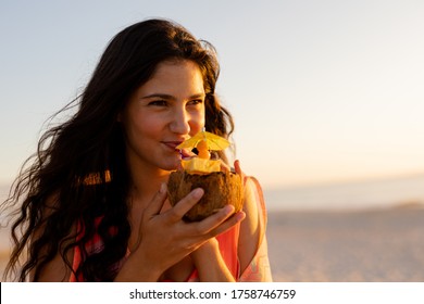 Mixed race woman enjoying her time at a beach with her freinds during sunset, drinking a drink from a coconut, looking away and smiling