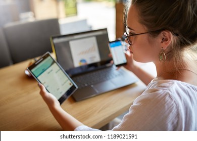 Mixed Race Woman In Glasses Working With Multiple Electronic Internet Devices. Freelancer Businesswoman Has Tablet And Cellphone In Hands And Laptop On Table With Charts On Screen. Multitasking Theme