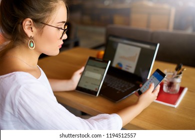 Mixed Race Woman In Glasses Working With Multiple Electronic Internet Devices. Freelancer Businesswoman Has Tablet And Cellphone In Hands And Laptop On Table With Charts On Screen. Multitasking Theme