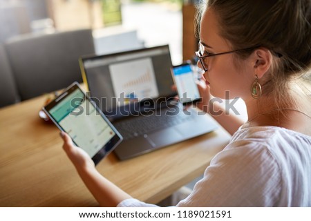 Mixed race woman in glasses working with multiple electronic internet devices. Freelancer businesswoman has tablet and cellphone in hands and laptop on table with charts on screen. Multitasking theme