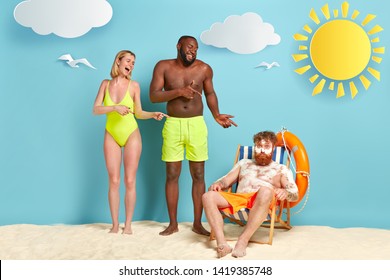 Mixed Race Woman And Man Laugh At Funny Friend Covered With White Burn Cream, Has Sunburnt Skin After Lying In Sun For Long Time, Sits At Beach Chair. Red Haired Male Applies Sunscreen On Skin