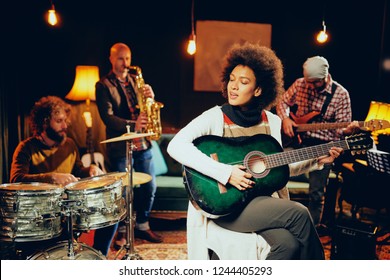Mixed race woman playing acoustic guitar and sitting with legs crossed in home studio. In background the rest of the band playing drums, saxophone and bass guitar. - Powered by Shutterstock