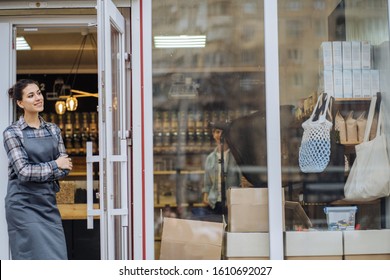 Mixed Race Woman Worker Or Store Owner With Standing In The Doorway Of Her Grocery Shop Looking Aside Smiling. Portrait Of Girl Assistant Wearing Apron Welcoming Inviting Open Door Of Store.