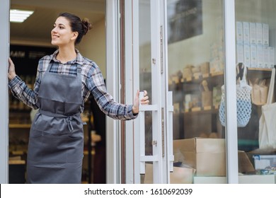 Mixed Race Woman Worker Or Store Owner With Standing In The Doorway Of Her Grocery Shop Looking Aside Smiling. Portrait Of Girl Assistant Wearing Apron Welcoming Inviting Open Door Of Store.