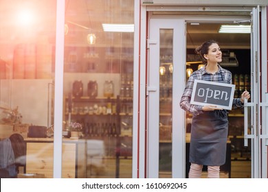 Mixed Race Woman Worker Or Store Owner With Standing In The Doorway Of Her Grocery Shop Looking Aside Smiling. Portrait Of Girl Assistant Wearing Apron Welcoming Inviting Open Door Of Store.