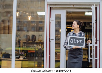 Mixed Race Woman Worker Or Store Owner With Standing In The Doorway Of Her Grocery Shop Looking Aside Smiling. Portrait Of Girl Assistant Wearing Apron Welcoming Inviting Open Door Of Store.